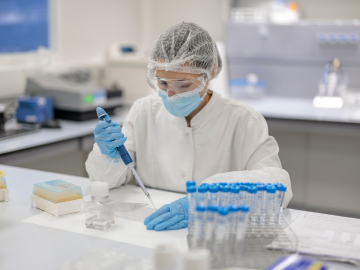 A researcher in a laboratory wearing a white coat, gloves, a face mask, and a protective hairnet, using a pipette to transfer liquid into a container. The workstation has test tubes, laboratory tools, and other scientific materials."