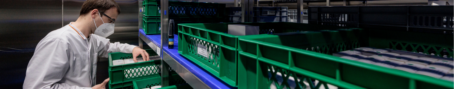 Close up of a  laboratory technician wearing a face mask and protective goggles inside a Eurofins storage and stability room, placing a green box with materials onto the shelf.