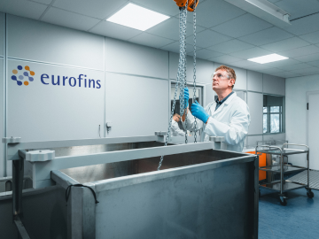 A scientist wearing a white lab coat, protective goggles, and blue gloves conducting Container Closure Integrity Testing showcasing advanced equipment and expertise in BioPharma Product Testing within a high-precision research environment.