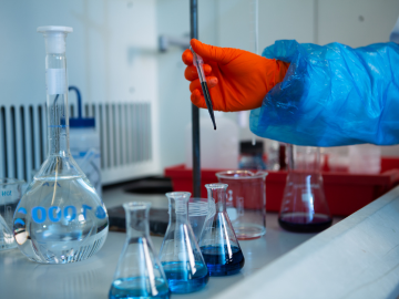 Close up of a researcher’s hand wearing safety goggles and lab coat. They are carefully conducting water analysis, expertly handling a pipette over lab flasks filled with blue liquid in a laboratory setting.