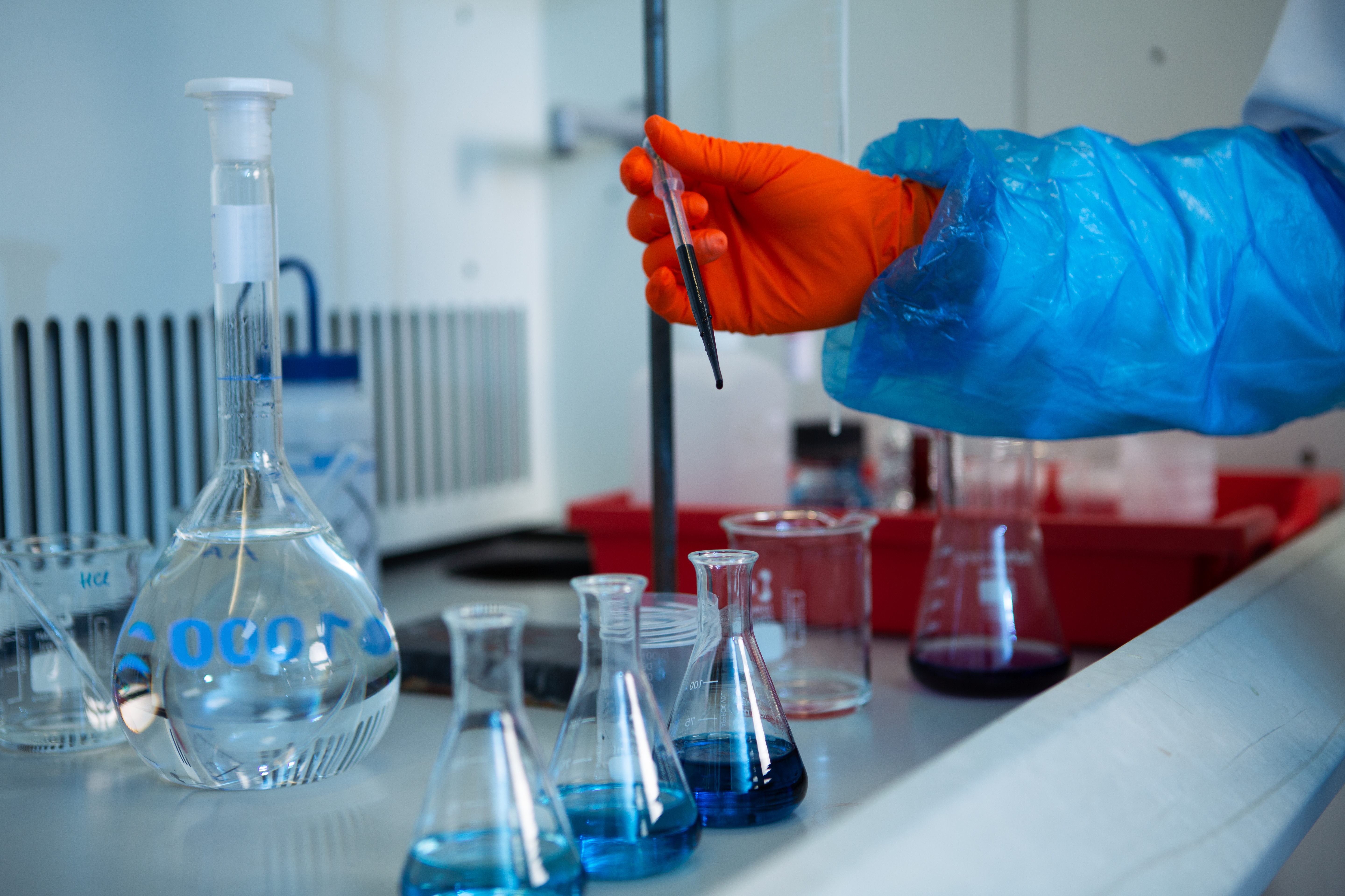 Close up of a researcher’s hand wearing safety goggles and lab coat. They are carefully conducting water analysis, expertly handling a pipette over lab flasks filled with blue liquid in a laboratory setting.