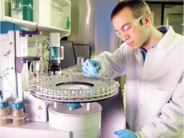 A laboratory technician wearing a white lab coat, gloves, and safety goggles, working with a Karl Fischer titration system. The technician is carefully placing or inspecting vials in a circular autosampler tray, surrounded by laboratory equipment and glassware.