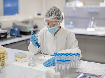 A researcher in a laboratory wearing a white coat, gloves, a face mask, and a protective hairnet, using a pipette to transfer liquid into a container. The workstation has test tubes, laboratory tools, and other scientific materials.