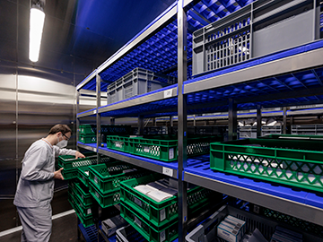 A man wearing a face mask and protective goggles inside a Eurofins storage and stability room, placing a green box with materials onto a shelf.