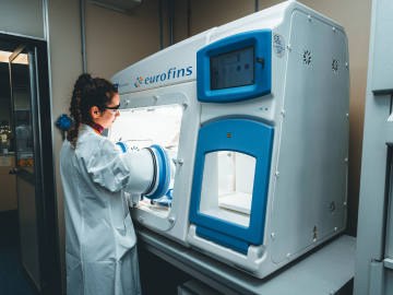 A scientist wearing a lab coat and protective glasses works in a laboratory. She is using a single advanced machine to conduct sterile and non-sterile microbiology testing. The environment is clean and professional, highlighting the focus on precision and safety in scientific research.