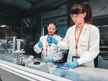 Two female scientists in a laboratory wearing safety goggles and lab coats, working with pipettes and medical devices. The image is related to medical device services, emphasizing precision and expertise in a clinical or research setting