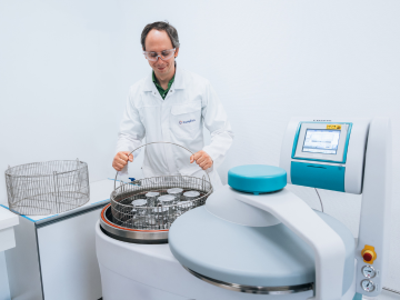 A laboratory technician wearing a white coat operates an autoclave during a disinfectant validation process. The scene shows a clean and sterile environment, with the technician handling a metal basket and the autoclave control panel visible in the foreground.