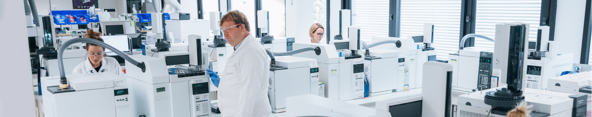 A modern laboratory equipped with advanced chromatographic instruments, where multiple scientists in white coats conduct analyses. The environment is clean and organized, with various devices used for identity, content, and purity determination.